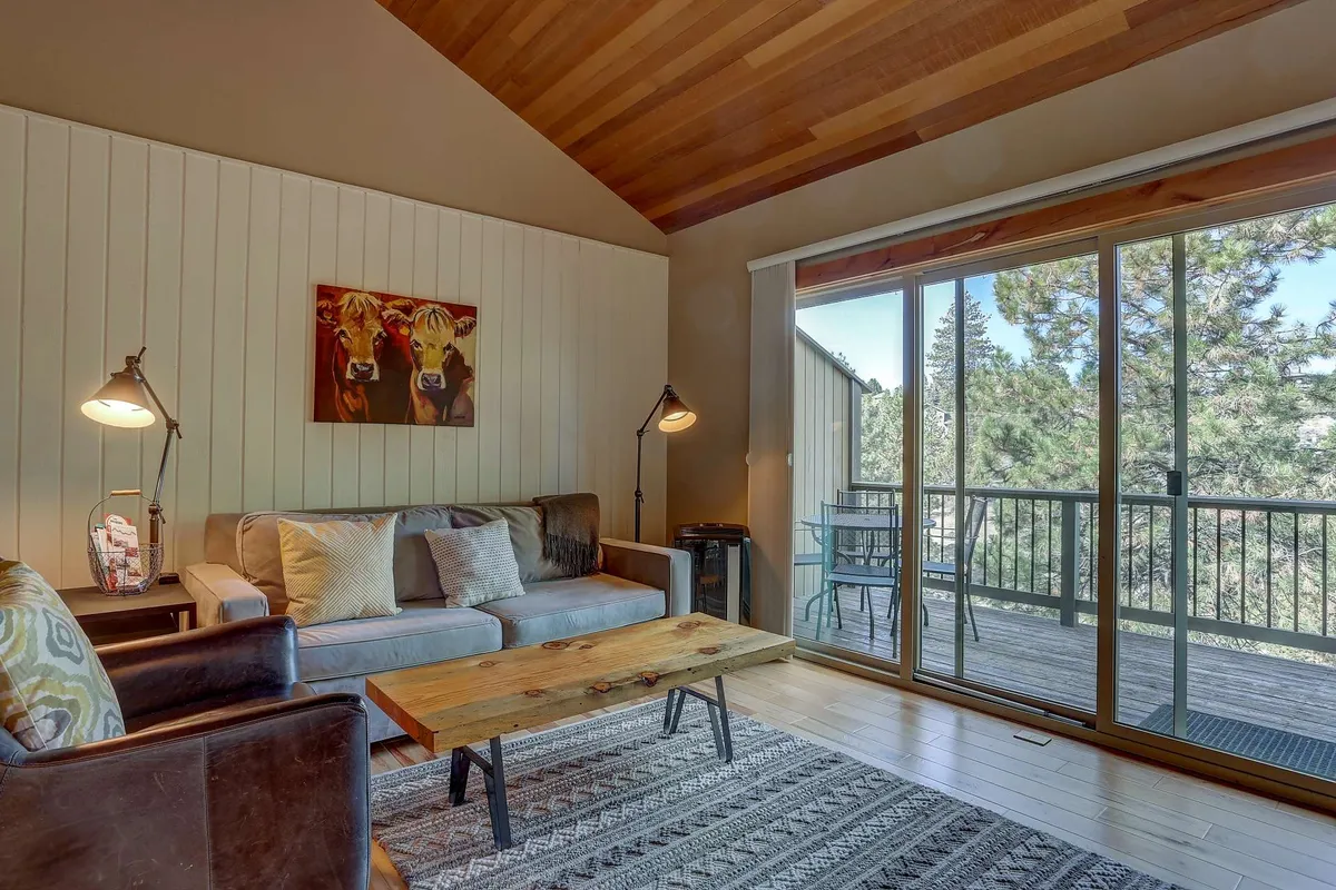 This inviting interior features a plush sofa with soft cushions, a coffee table perfect for board games or drinks, and large glass doors that open to a private deck. Artistic touches, such as cow-themed artwork, add charm, while ample sunlight creates a cheerful atmosphere for both relaxation and socializing.