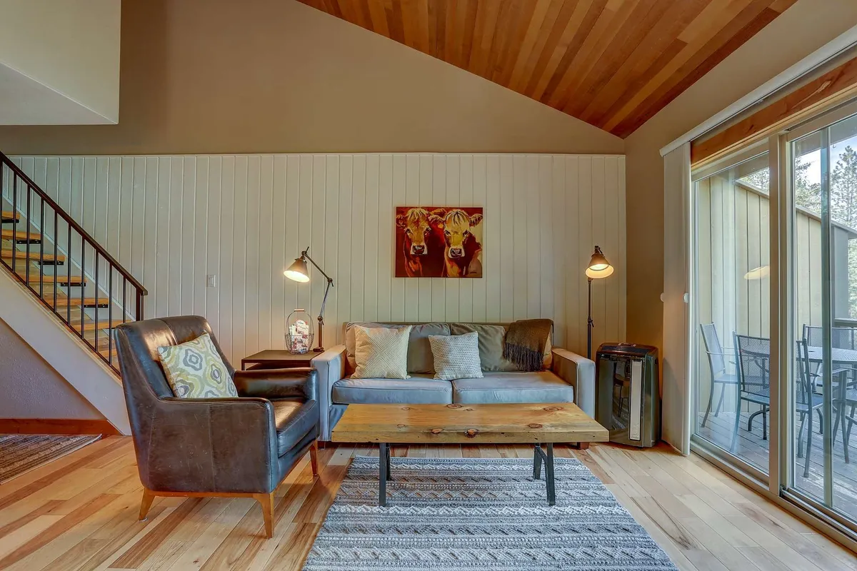 A cozy living room with a sofa adorned with decorative cushions, a wooden coffee table, art on the wall, a floor lamp, and a large sliding glass door. The room is bathed in natural light, suggesting comfort and a welcoming environment for all guests.
