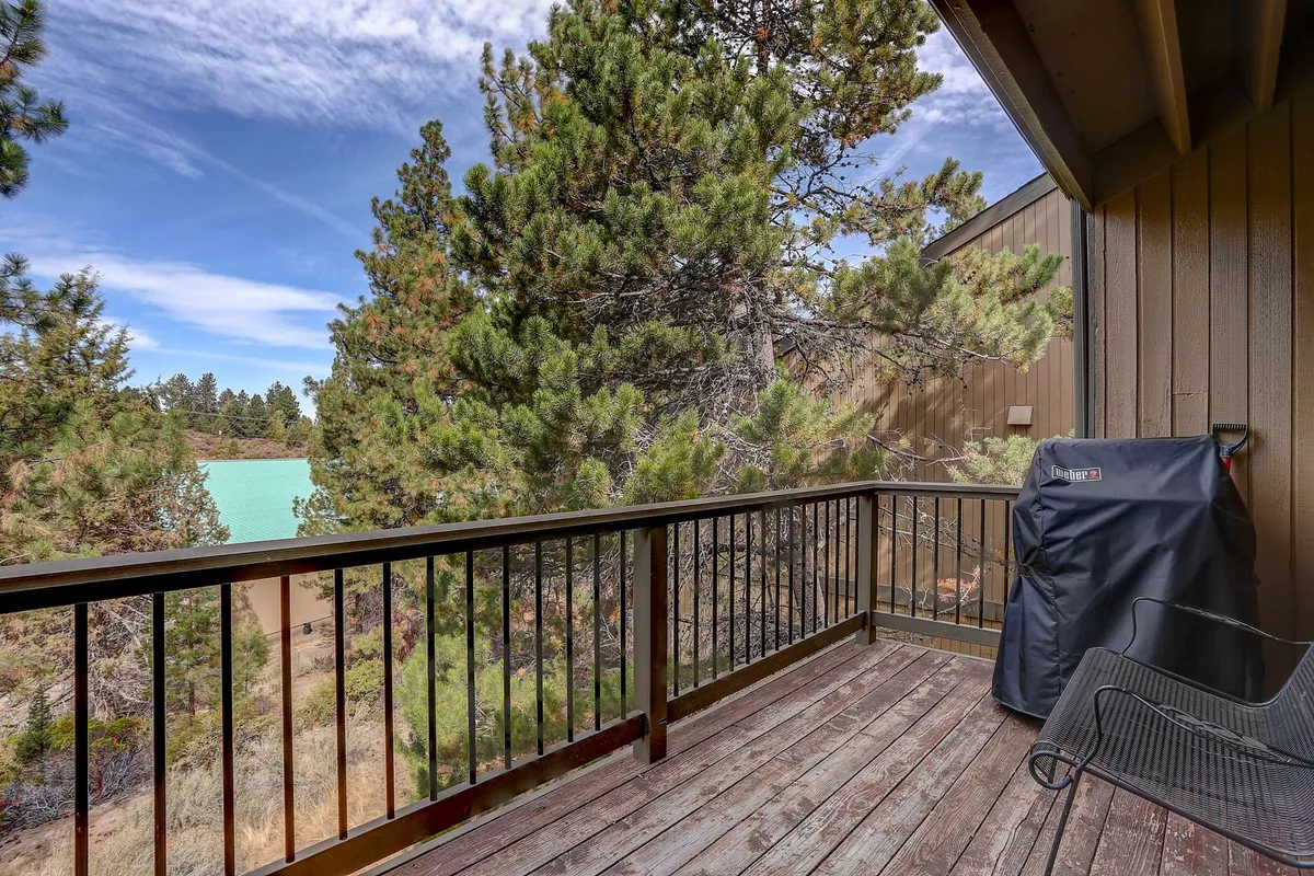 A serene wooden balcony overlooks lush green trees and a shimmering stretch of blue water. The space features ample room for relaxing, enjoying the view, or barbecuing, with the natural forest setting visible from the deck.