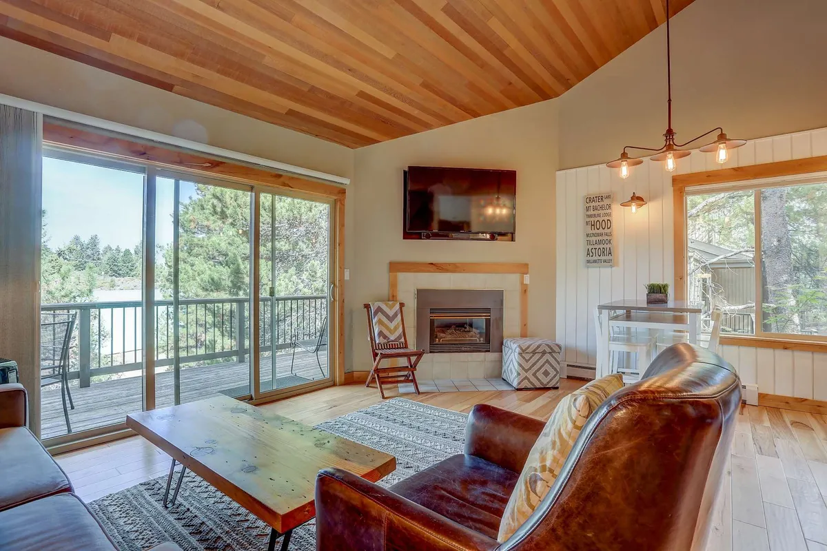 This interior image showcases a welcoming living room with a gas fireplace as the focal point, flanked by large windows that let in abundant natural light and offer tranquil forest views. The space features comfortable, plush seating arranged around a coffee table, perfect for relaxation or conversation. The vaulted wooden ceiling enhances the room's sense of spaciousness and warmth.