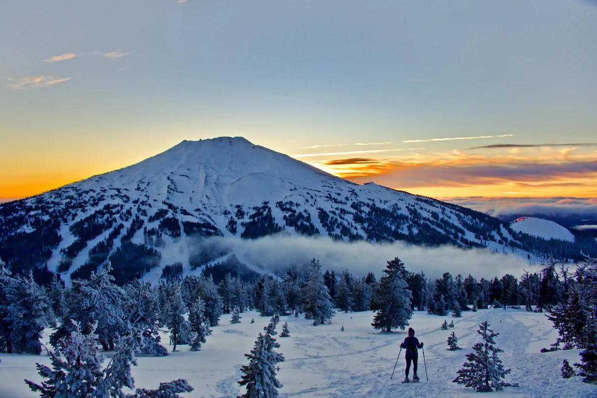 A snow-capped mountain under a vibrant sunset sky, with a skier descending through fresh powder surrounded by towering pine trees. The scene radiates the exhilaration and beauty of Mt. Bachelor in winter, capturing both the adventure and the majestic natural setting.