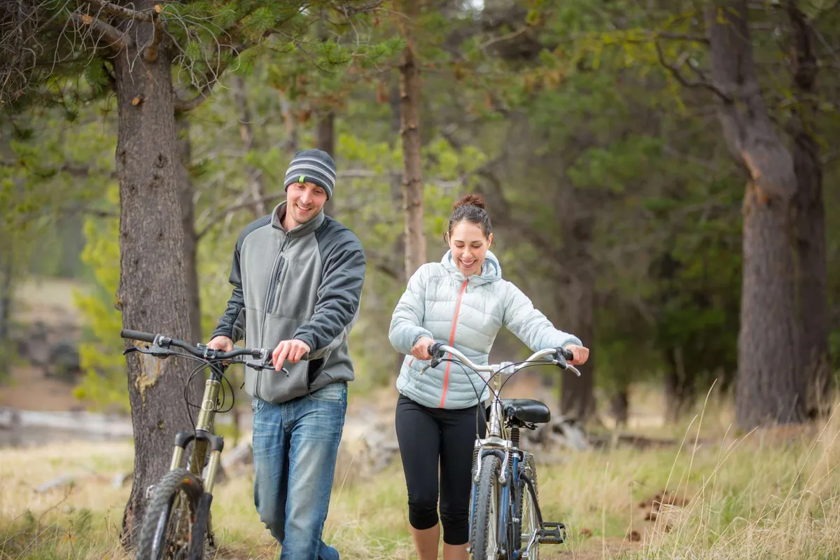 A couple strolls along a tree-lined path with bicycles, surrounded by a lush natural landscape. The scene evokes adventure, activity, and direct access to outdoor trails, with a joyful and relaxed mood.