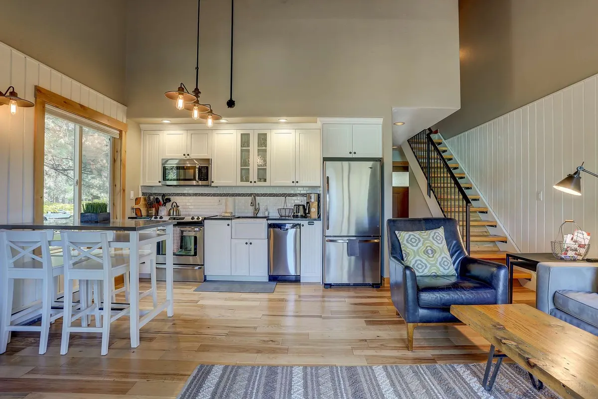 The image showcases a beautifully remodeled open-concept living area, featuring a spacious kitchen island perfect for cooking, comfortable seating ideal for relaxing or socializing, and a stylish staircase leading to the second level. The warm lighting and inviting décor create a cozy, welcoming ambiance that encourages guests to unwind after a day of mountain adventure.