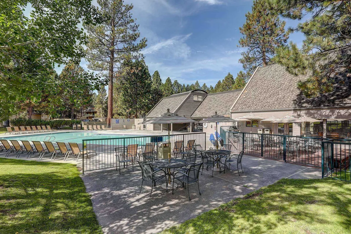 A spacious outdoor swimming pool is surrounded by comfortable lounge chairs and shaded seating areas, with a clubhouse in the background and tall pine trees providing a natural, resort-like setting. People are gathered around, enjoying the sun and relaxed atmosphere.