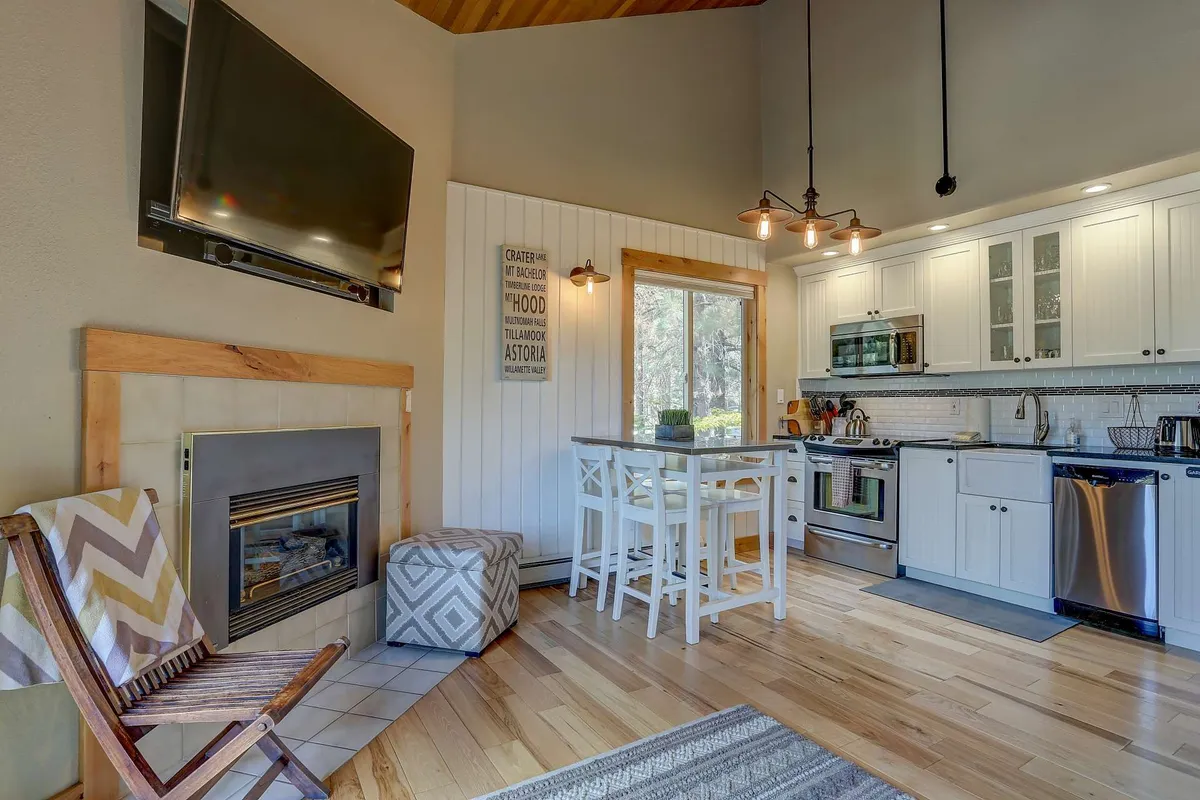 A bright, open-plan interior with a dining table set for meals, a visible kitchen area, and a fireplace that adds warmth. Sunlight pours in through wide windows, illuminating the dining and living areas, creating an inviting setting for shared meals and entertaining guests.
