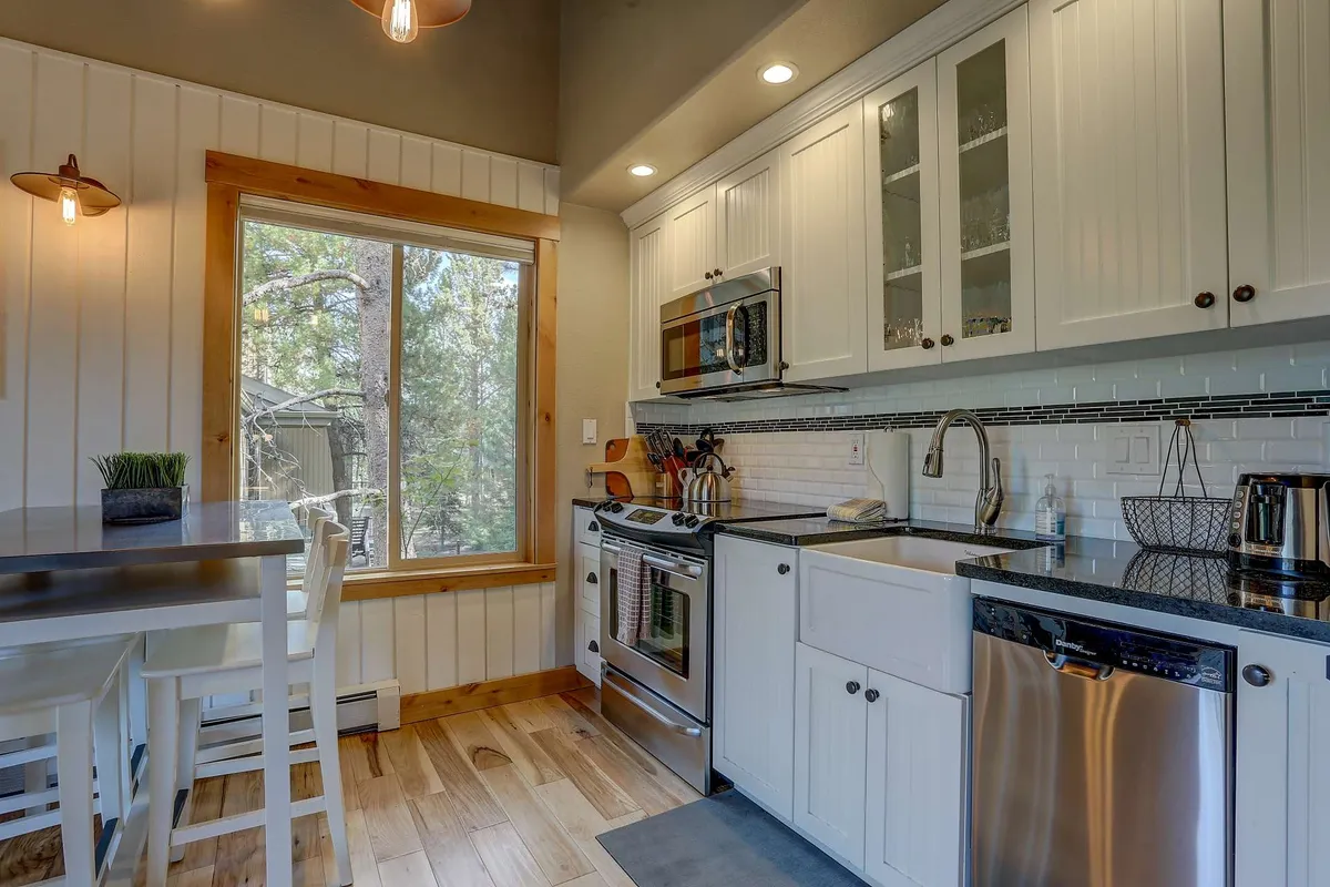 This image showcases a cozy kitchen and dining area with modern appliances, a large window overlooking trees, and a central dining table. The kitchen is equipped with stainless steel appliances and generous cabinetry, all bathed in natural light from the window.