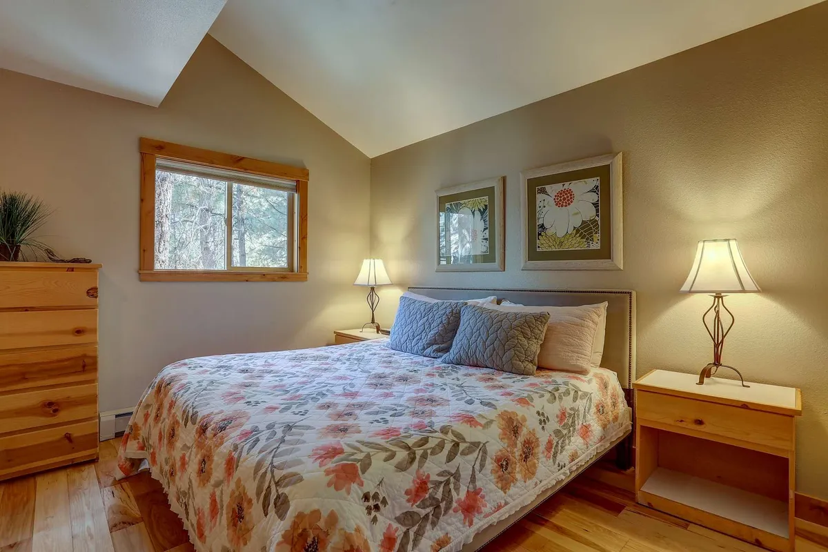 A tranquil king or queen bedroom featuring a bed with a floral quilt, two bedside lamps casting warm light, framed wall art, and a window letting in natural light. The overall atmosphere is peaceful and inviting, with subtle decor elements enhancing the restful mood.
