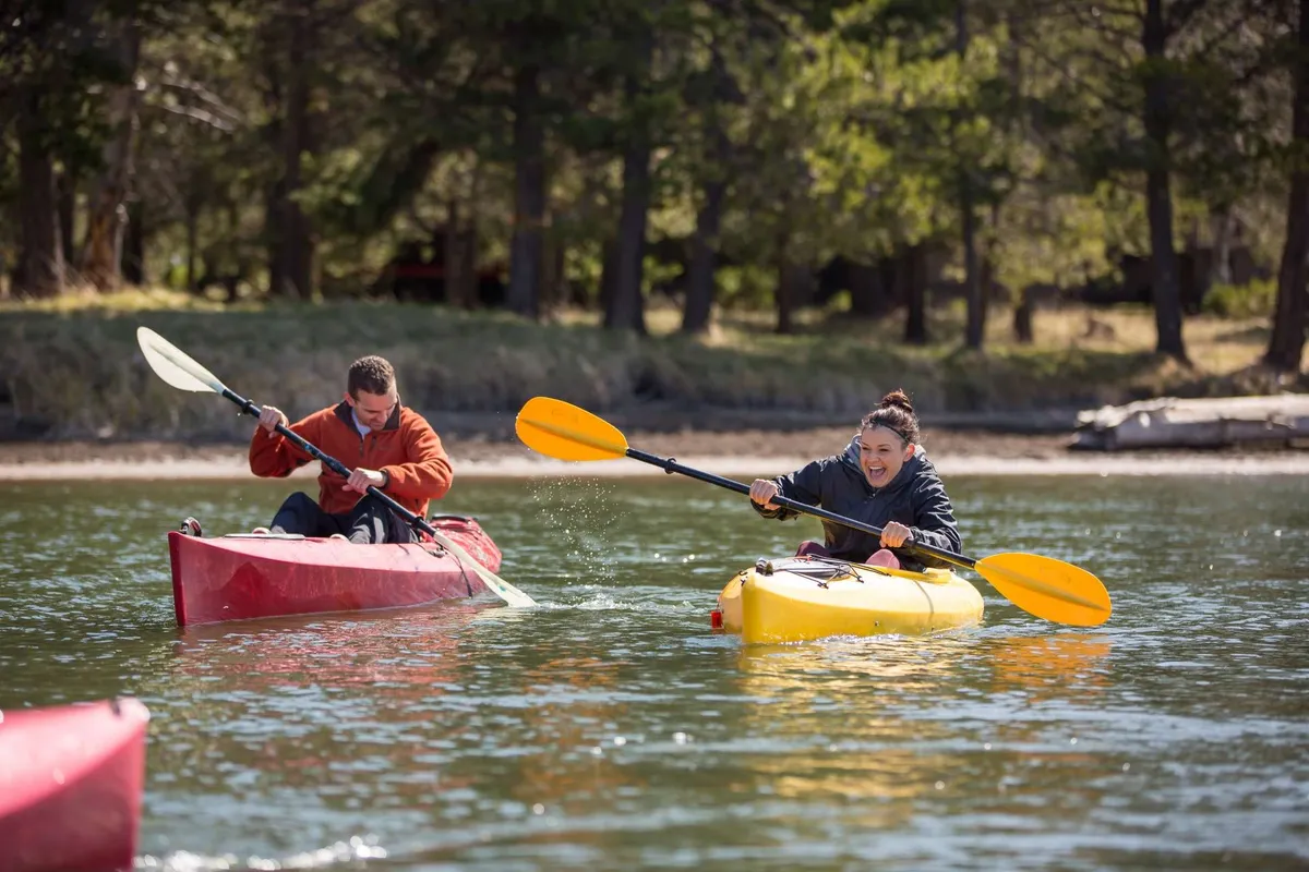 Two kayakers paddle in brightly colored kayaks across calm, reflective water, surrounded by lush greenery. The energy of their movement and the natural beauty of the setting evoke the fun and adventure of summer activities.