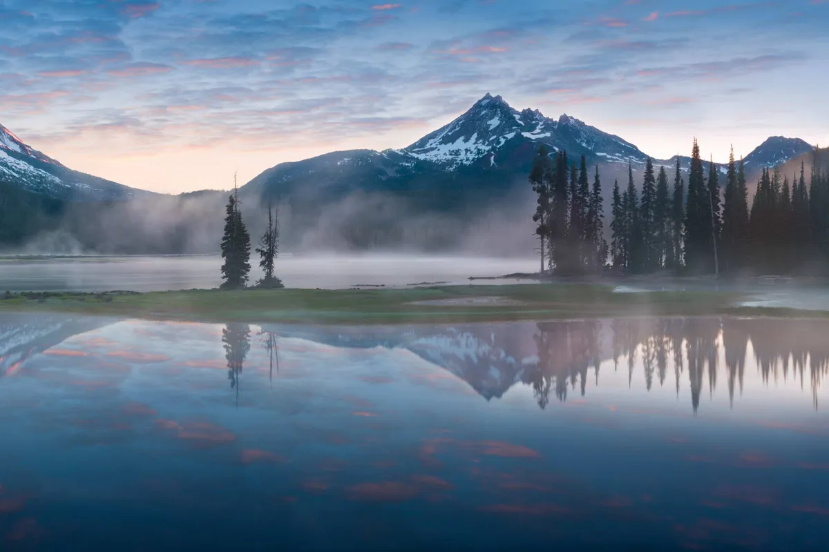 A serene landscape featuring a tranquil mountain lake surrounded by trees, with distant peaks reflected in the water. The scene evokes calm and the beauty of nature, hinting at outdoor exploration and moments of quiet reflection.